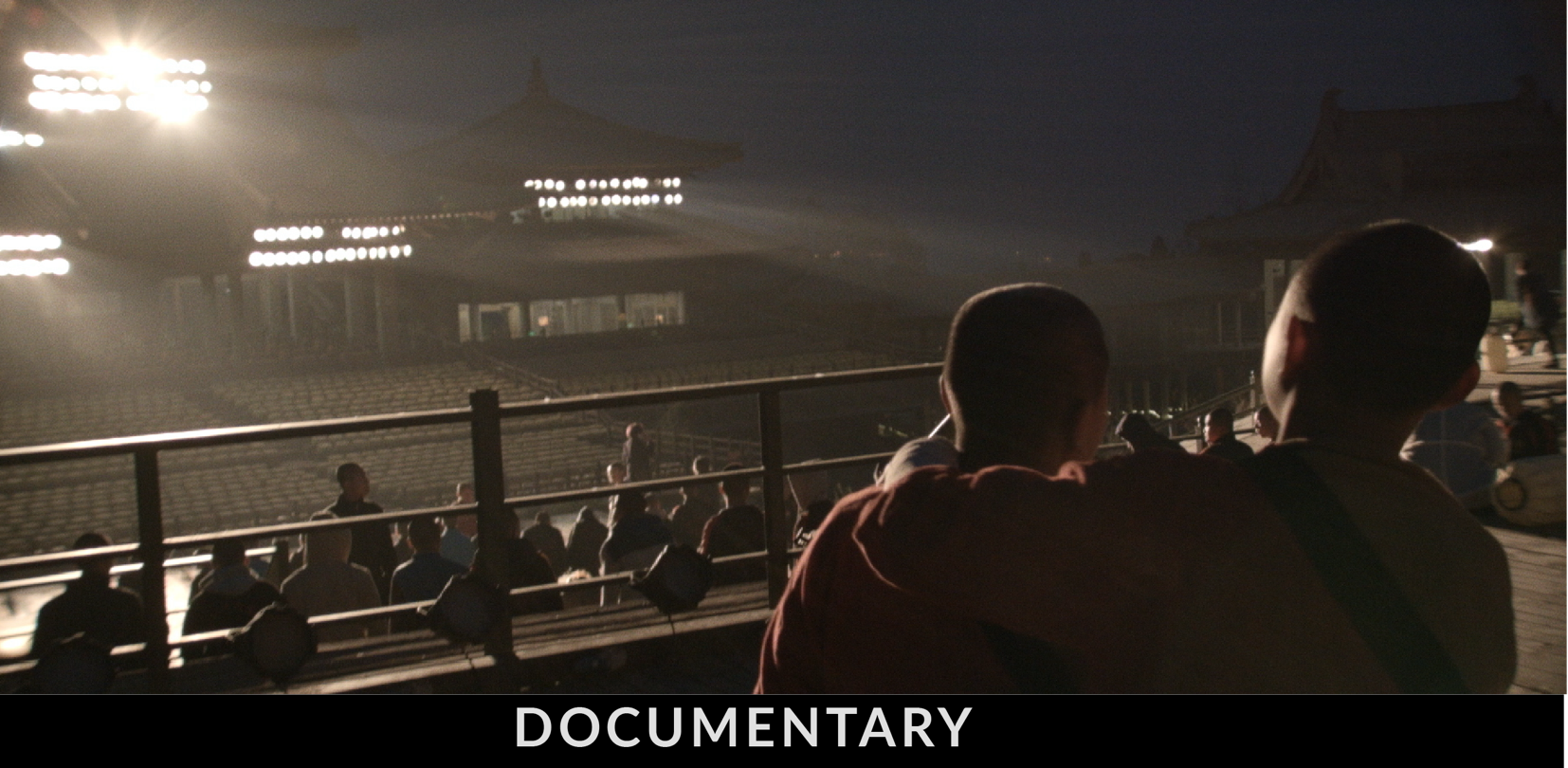 Two young boys in robes stand on a stage for a kungfu show, facing a lit-up traditional building with a group of people sitting on the ground in the background.