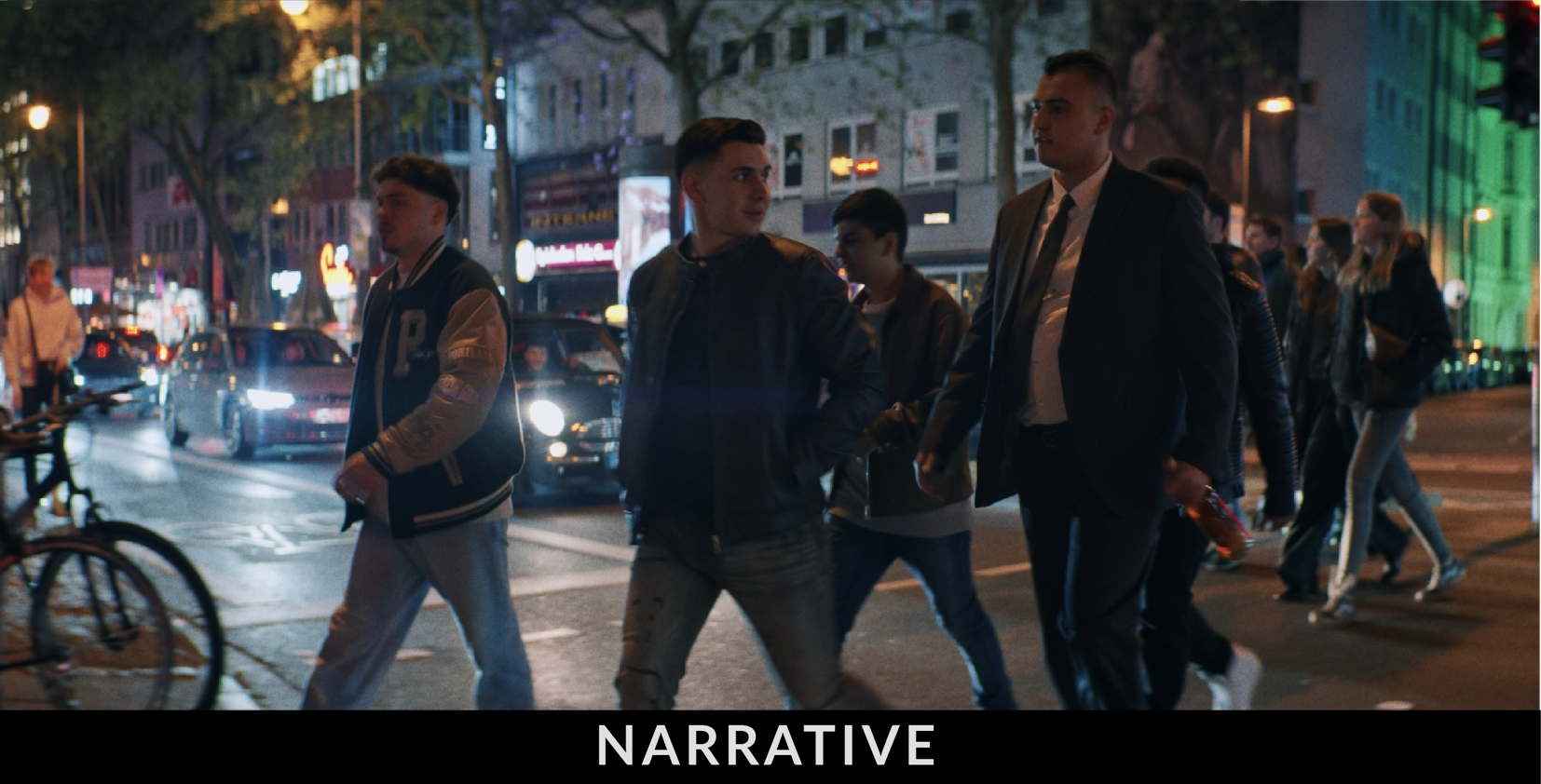 A group of people walking on a city street at night, with cars and bicycles nearby, and buildings illuminated in the background.